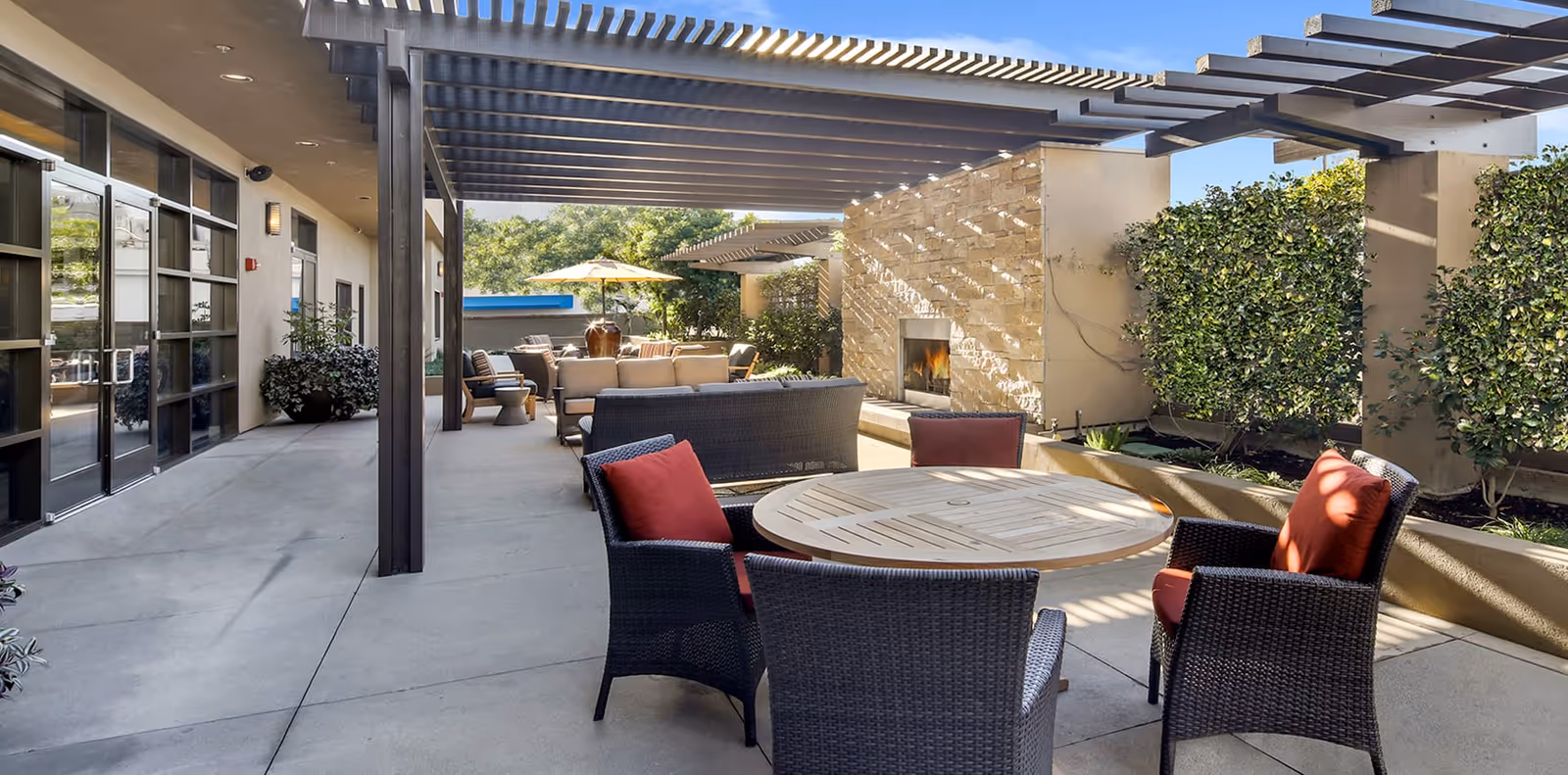 Outdoor patio area with a round wooden table surrounded by four wicker chairs with red cushions. There is a pergola overhead providing partial shade, a stone wall with a built-in fireplace, and additional seating with sofas and an umbrella in the background. Green shrubs line the right side of the patio.