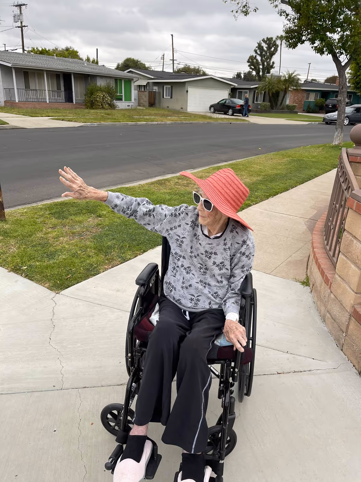 An elderly woman in a wheelchair wearing a red sunhat, white sunglasses, a gray floral sweater, black pants, and pink slippers is sitting on a sidewalk next to a grassy area. She is raising her left arm and looking in that direction. In the background, there are single-story houses, a street, and a cloudy sky.
