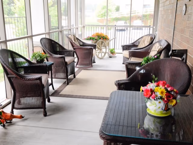 A screened-in porch with several dark wicker chairs arranged around a beige area rug. There are small tables with colorful flower arrangements placed on them, and a decorative cart with flowers is visible at the far end. The porch overlooks a green outdoor area.