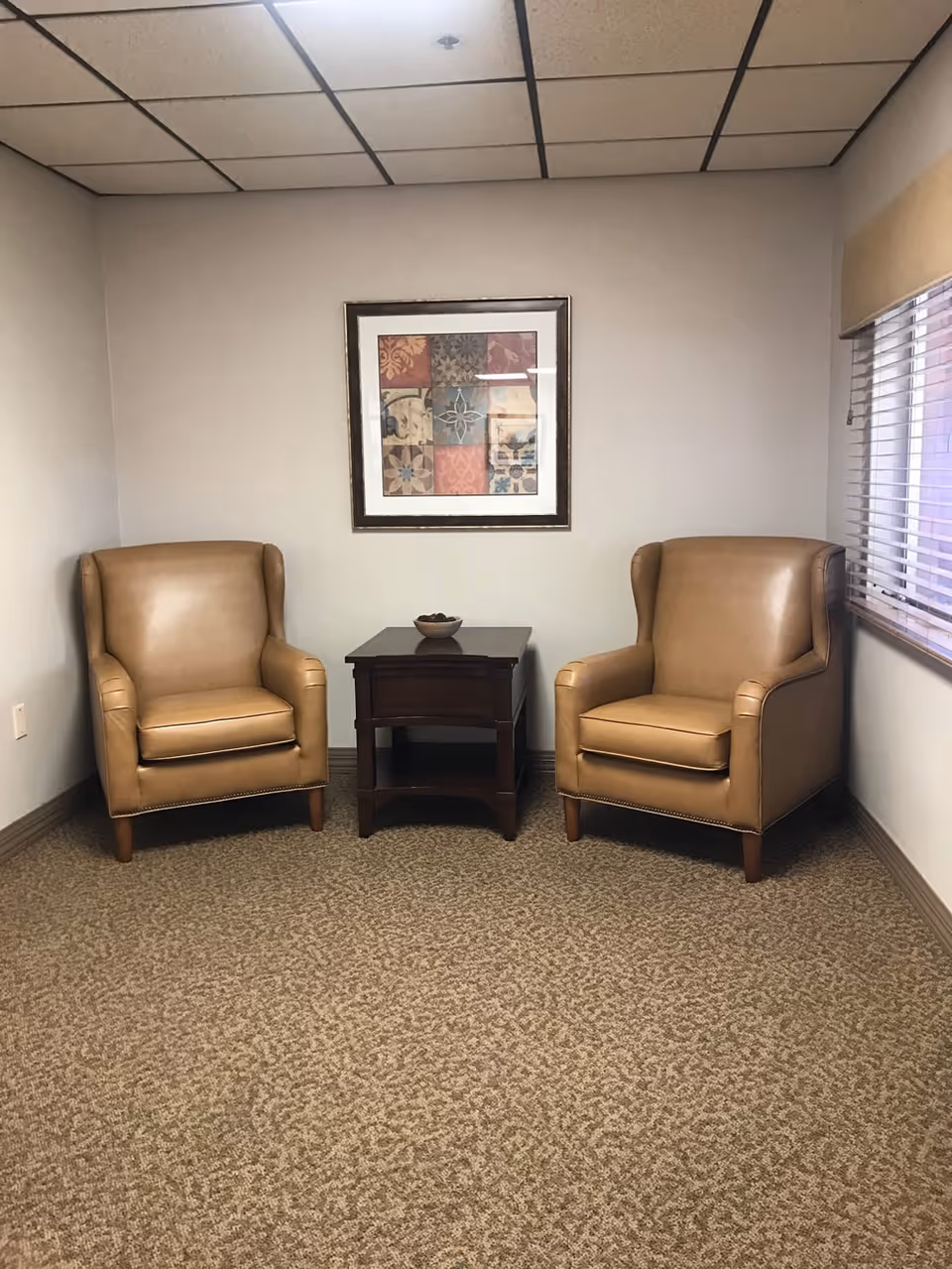 A small sitting area with two tan leather armchairs positioned on either side of a dark wooden side table with a bowl on top. The walls are light-colored, and there is a framed artwork with a patchwork design hanging above the table. A window with blinds is visible on the right side of the image. The floor is carpeted with a patterned design.