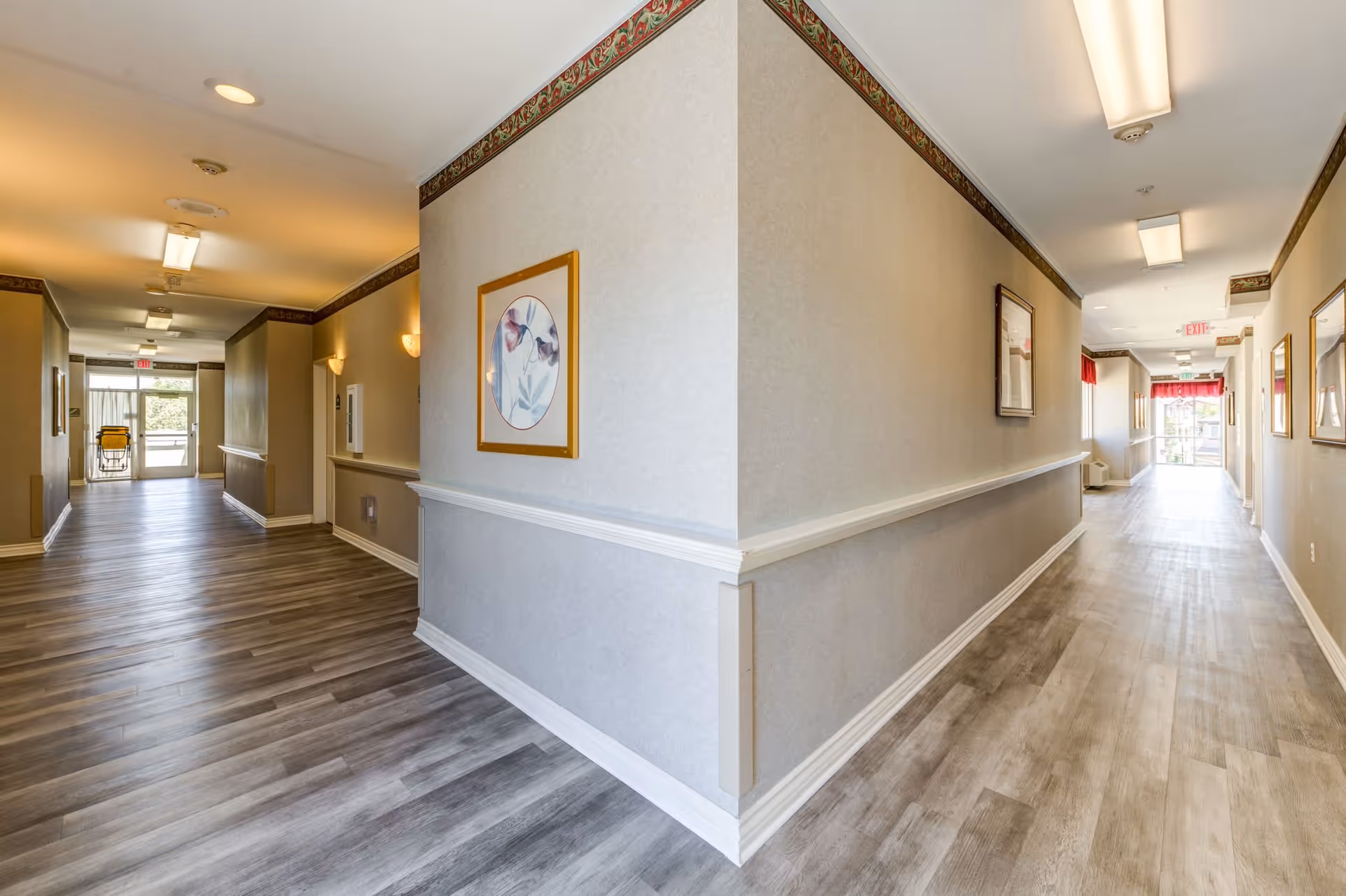 Well-lit interior hallway in a senior living facility with wood-look flooring, handrails, and framed artwork leading to exits at both ends.