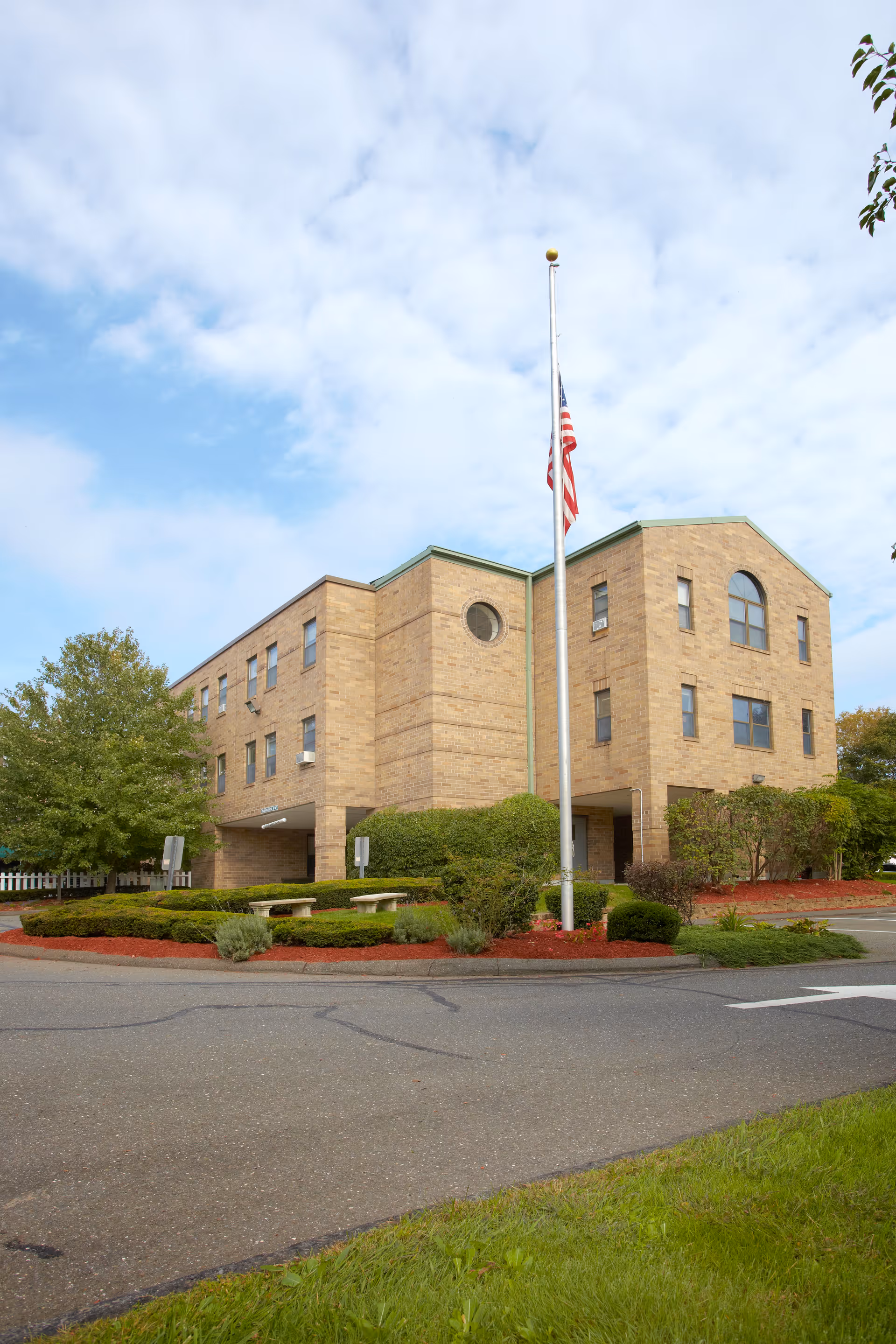 Exterior view of a three-story brick building with multiple windows, surrounded by landscaped bushes and trees. An American flag is flying at half-mast on a flagpole in front of the building under a partly cloudy sky.