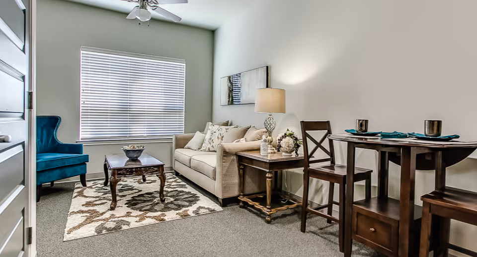 Small furnished living room with a beige sofa, teal armchair, coffee table, side table with lamp, and a small dining table.