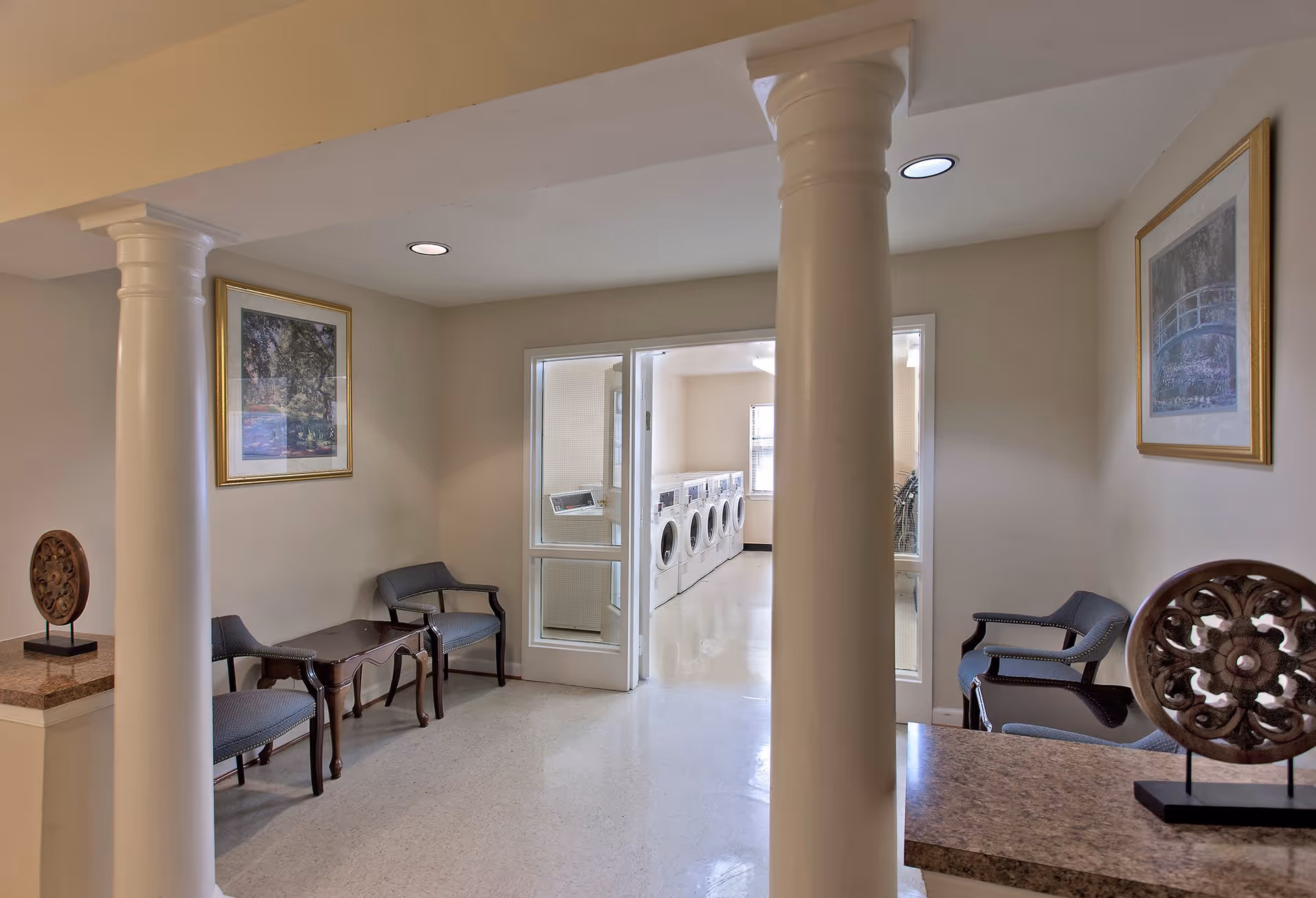 Interior view of a senior living facility hallway with two white columns, seating area with chairs and a small table, decorative wall art, and a laundry room with washing machines visible through glass doors.
