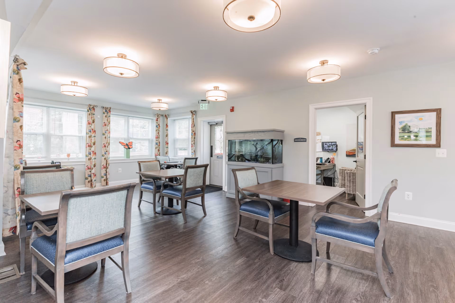 Bright communal dining room with multiple tables and chairs, large windows with floral curtains, and a fish tank against the wall.