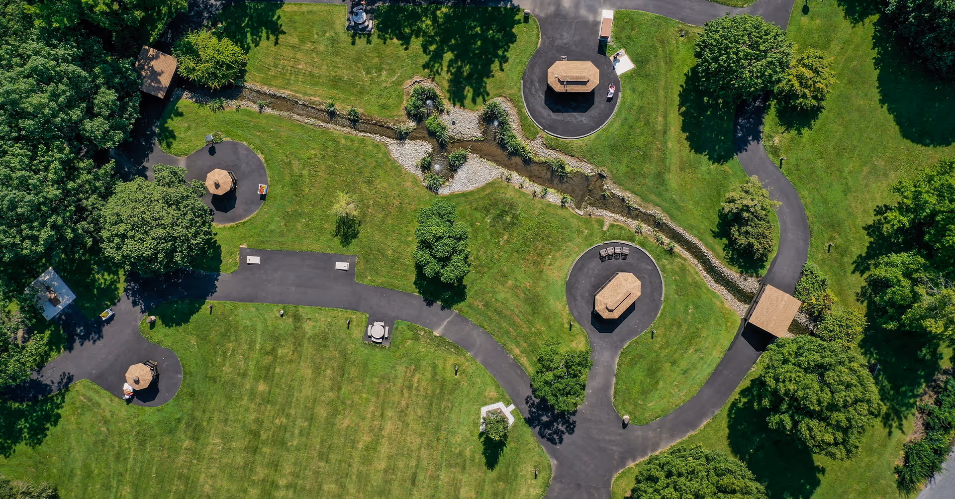 Aerial view of a green outdoor park area with paved walking paths, several picnic tables with umbrellas, a small stream running through the center, and scattered trees providing shade.