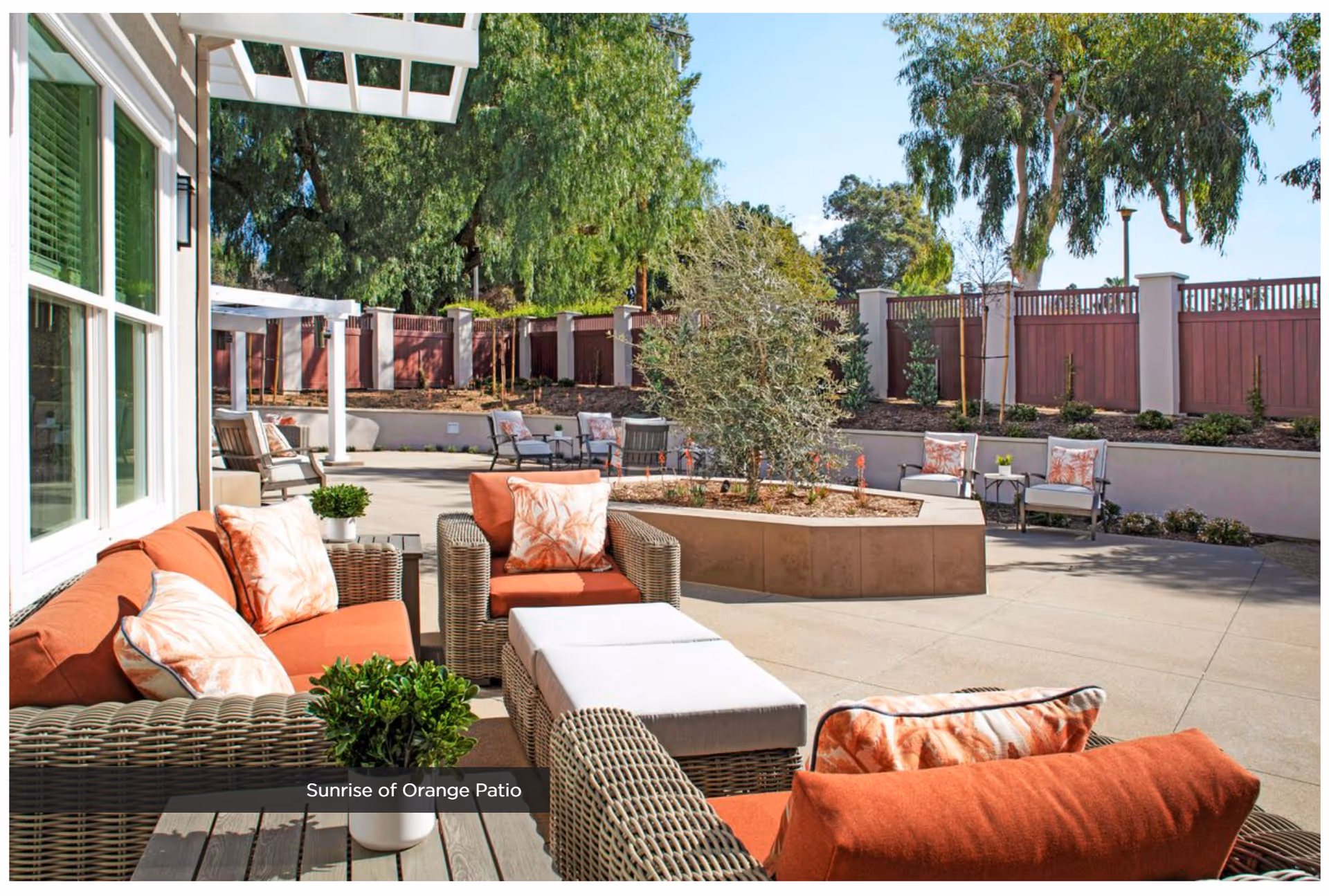 Outdoor patio area at Sunrise Of Orange featuring wicker furniture with orange cushions and patterned pillows, a central planter with a small tree, additional seating with chairs and small tables, surrounded by a wooden fence and lush green trees under a clear blue sky.