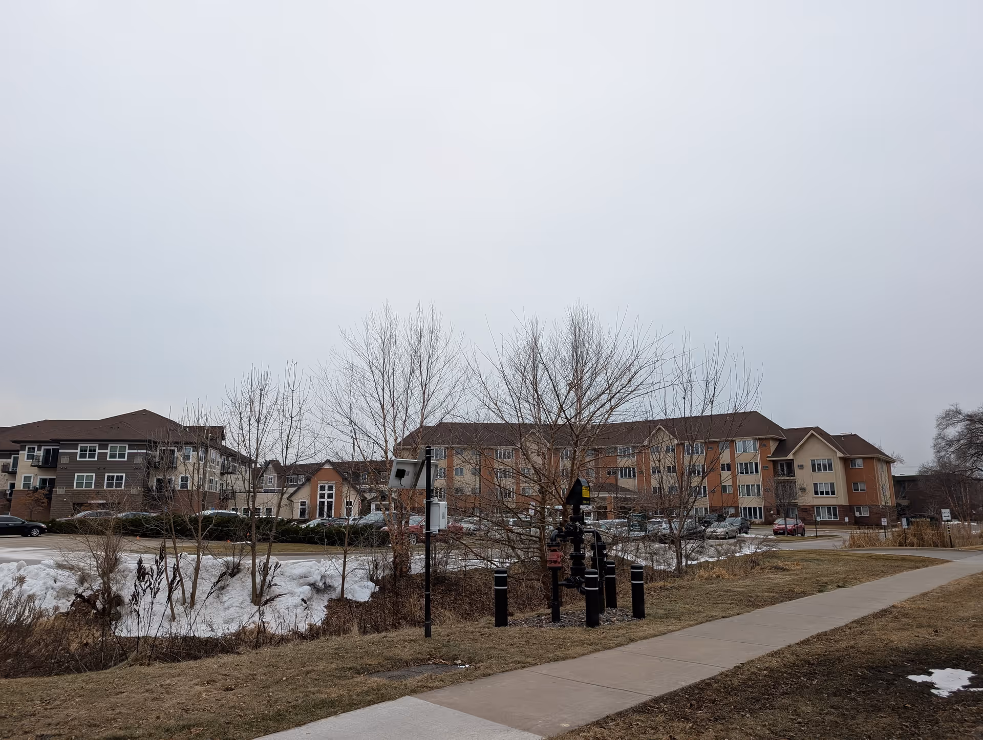 Front exterior view of a multi-story senior living building with a sidewalk and leafless trees in the foreground under an overcast sky.