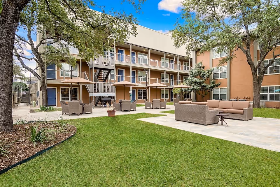 Outdoor courtyard area of a senior living facility with green grass, trees, and several seating arrangements including wicker chairs with umbrellas and a cushioned sofa set on a paved patio. The building surrounding the courtyard is three stories high with balconies and staircases.