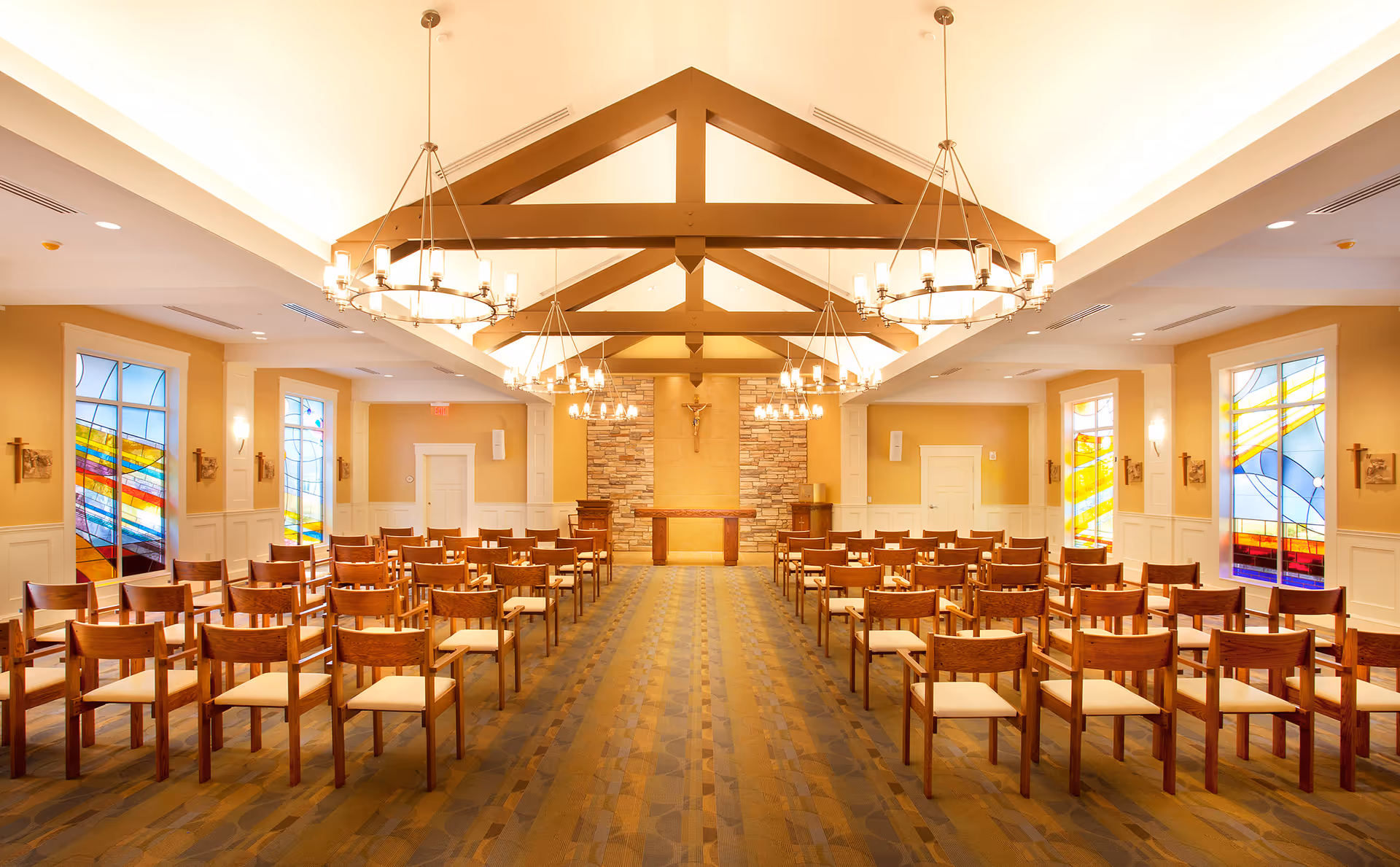 Sunlit chapel-like interior with rows of wooden chairs facing an altar beneath exposed ceiling beams and stained-glass windows.