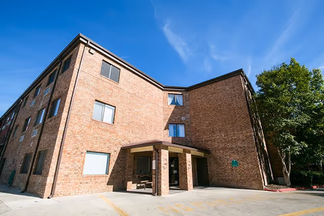 Exterior view of a three-story brick building with multiple windows and a covered entrance under a clear blue sky. There are trees on the right side and a paved area in front of the entrance.