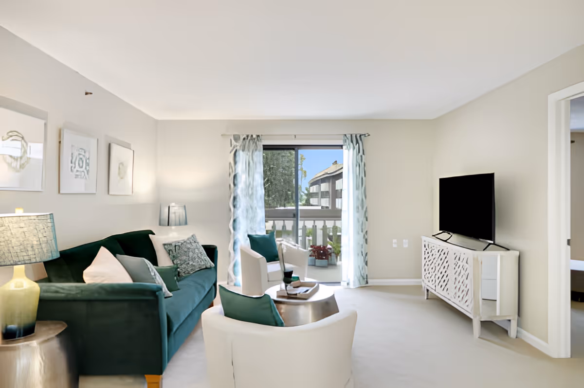 A bright living room with a green sofa, two white armchairs with teal cushions, a round coffee table, and a TV on a white console. There are three framed artworks on the wall above the sofa, a lamp on a side table, and sliding glass doors with patterned curtains leading to a balcony with plants and a view of another building.