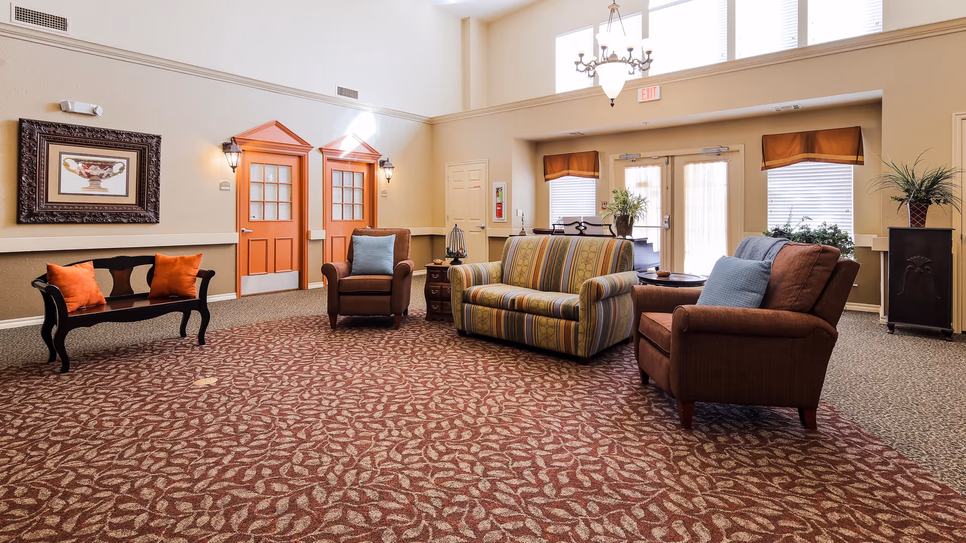 A spacious and well-lit living room area with patterned carpet, a striped sofa, two brown armchairs with blue cushions, a black bench with orange cushions, and a small wooden side table. The room features two orange doors with glass panels, large windows with blinds, a chandelier, and decorative plants.