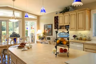 Bright kitchen and dining area with light wood cabinetry, a large island with fruit and decorative items, blue pendant lights, and a dining table set near large windows and a glass door leading outside.