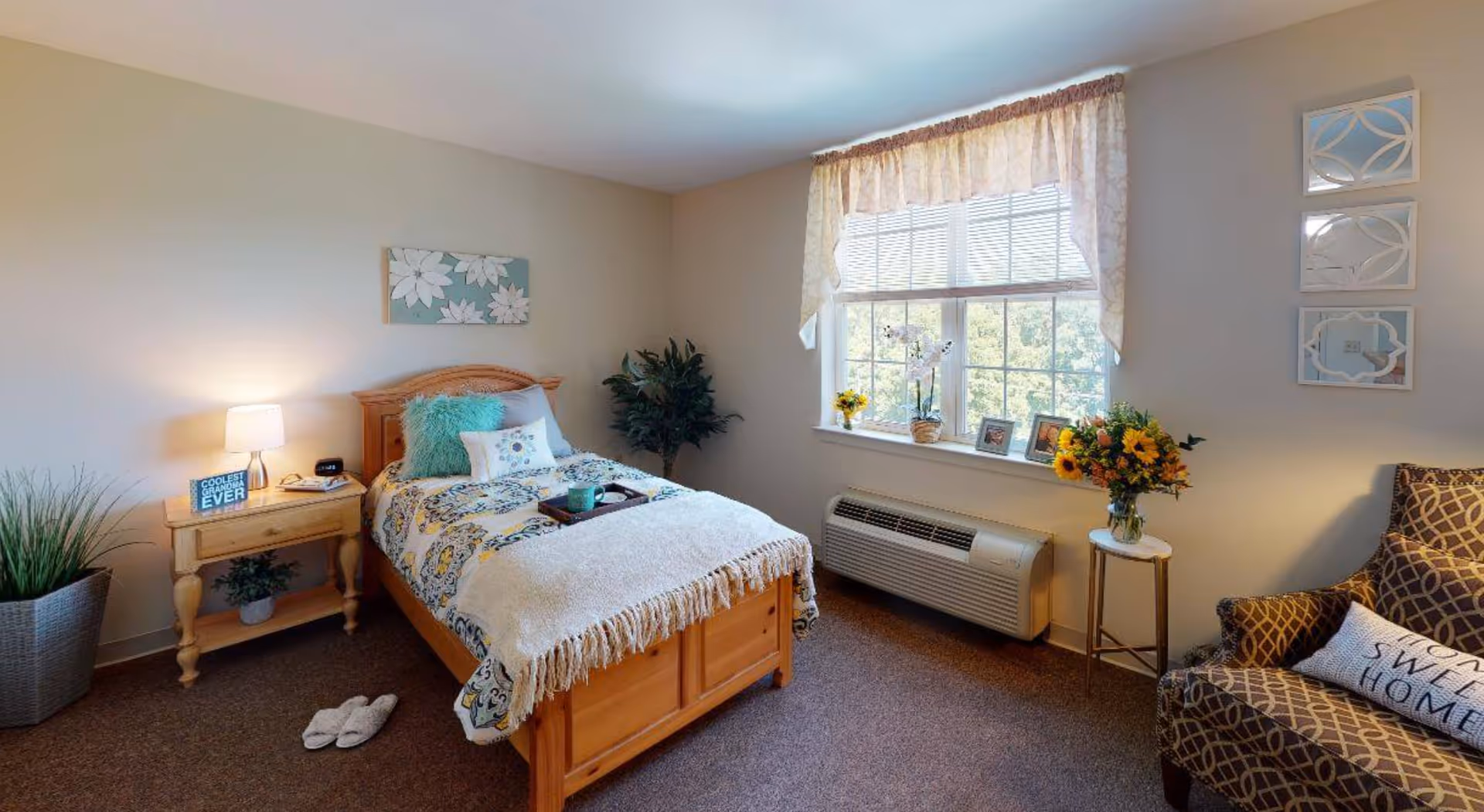 A cozy bedroom with a single wooden bed covered with a patterned quilt and pillows. A small wooden nightstand with a lamp, clock, and a sign that reads 'Coolest Grandma Ever' is beside the bed. There is a large window with sheer curtains letting in natural light. A patterned armchair with a pillow that says 'Home Sweet Home' is next to a small round table holding a vase of flowers. The room also features decorative wall art, a potted plant, and slippers on the carpeted floor.