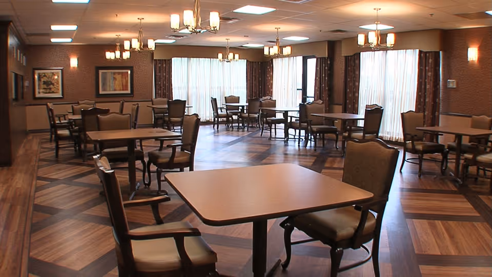 A spacious dining room with multiple square tables and cushioned chairs arranged neatly. The room features large windows with sheer curtains allowing natural light to fill the space, wooden patterned flooring, and warm lighting fixtures on the ceiling and walls. Framed artwork is visible on the walls.
