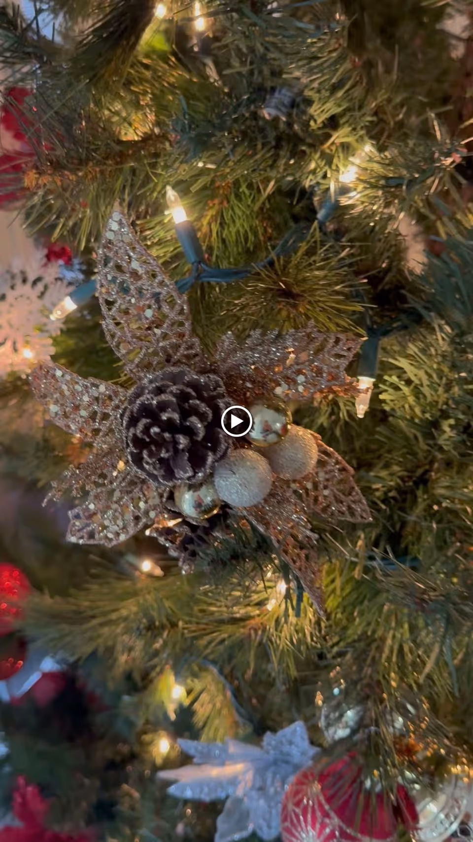 Close-up of a decorated Christmas tree branch with glowing string lights, a glittery poinsettia ornament, pinecone and gold baubles.