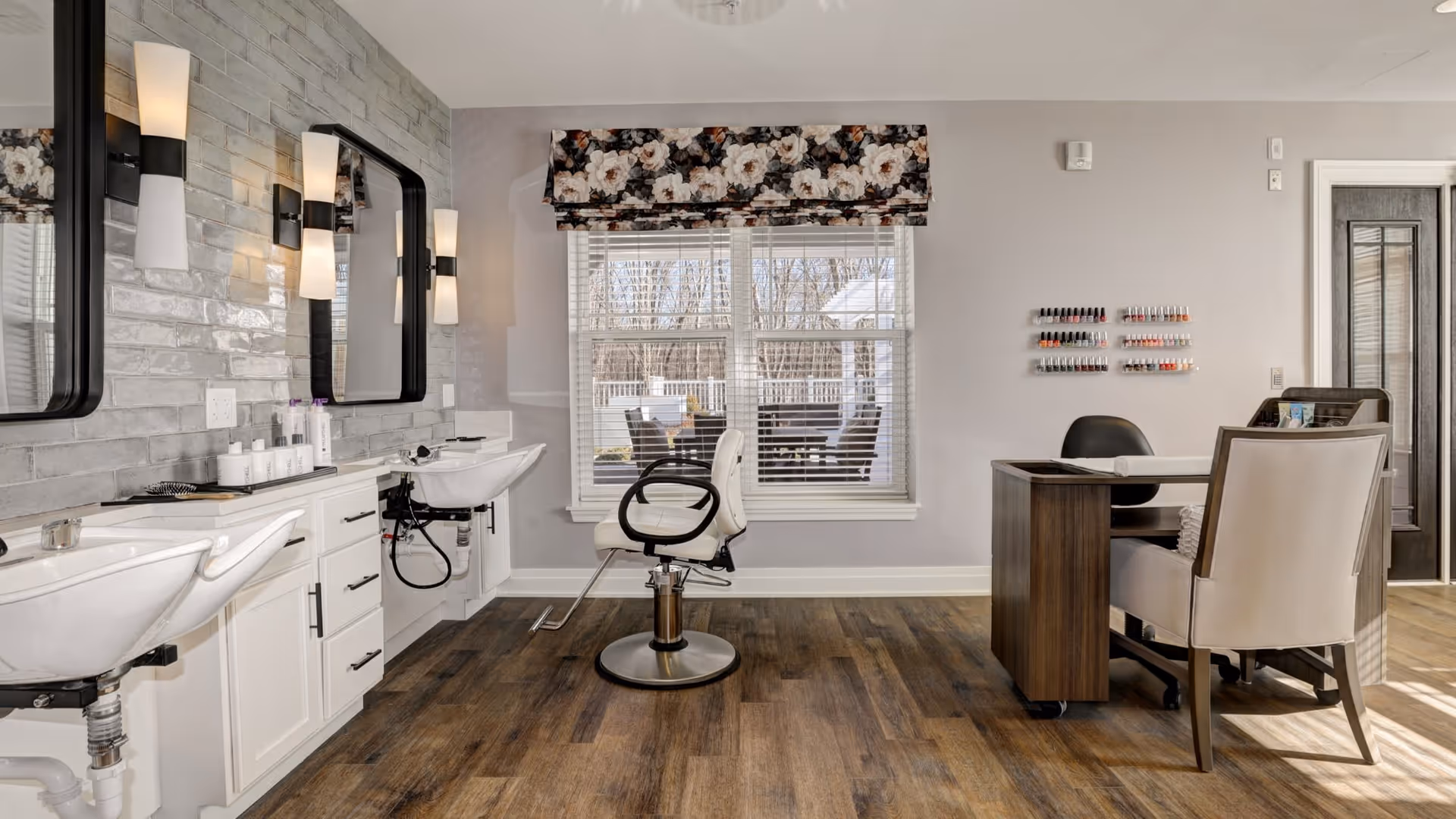 A bright and clean salon area inside a senior living facility featuring two white hair washing sinks with mirrors and wall lights above them on the left side. In the center, there is a white salon chair facing a large window with floral valance curtains. On the right side, there is a manicure station with a wooden desk, two chairs, and a wall-mounted display of various nail polish colors. The floor is wood-style, and the walls are painted light gray.