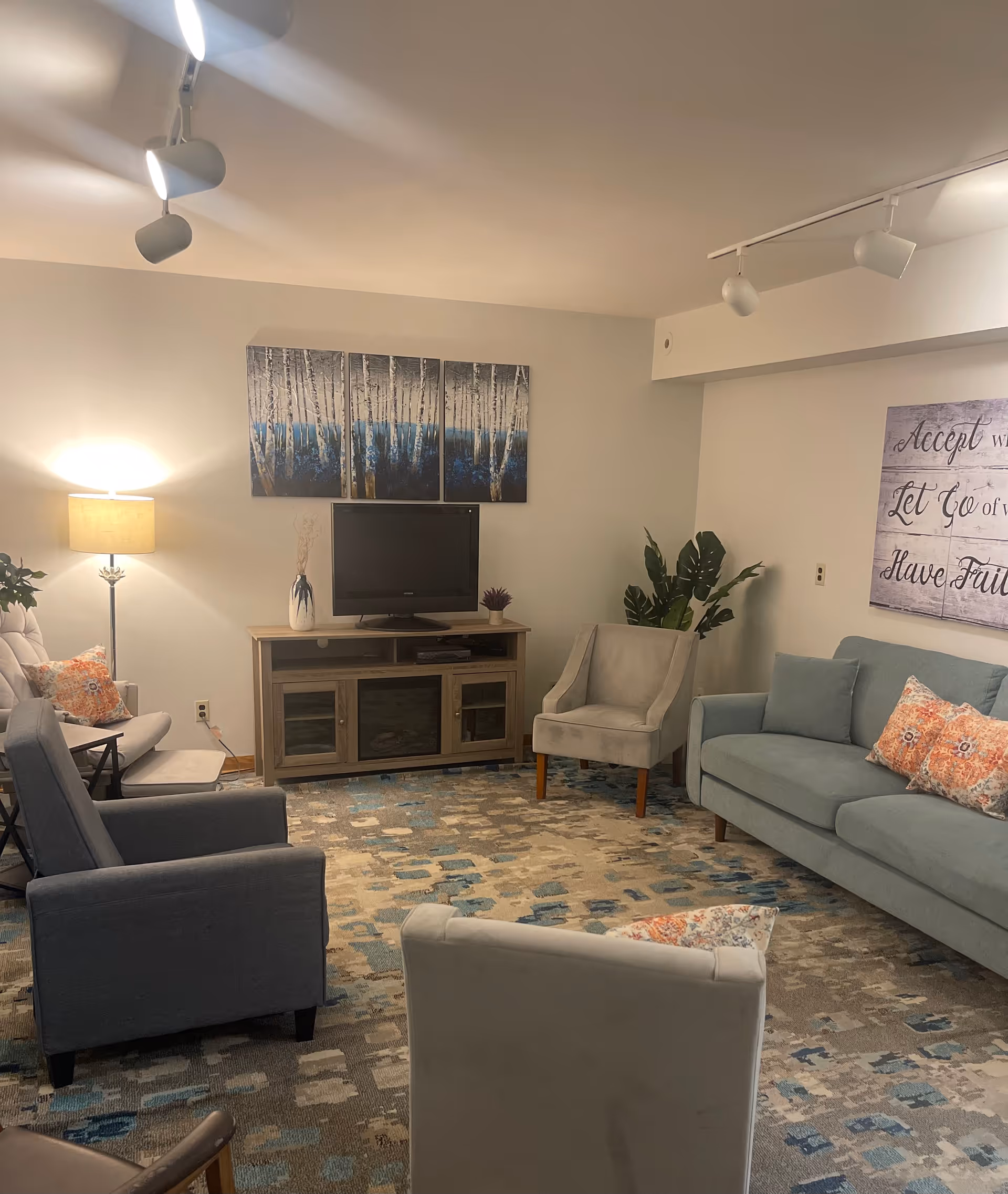 A cozy living room with a light blue sofa, two armchairs, a TV on a wooden stand, a floor lamp, and decorative wall art. The room has a patterned carpet and soft lighting.