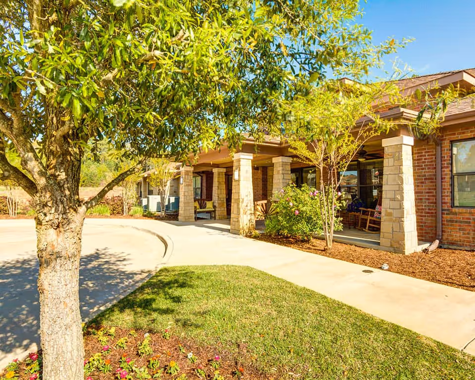 Front exterior view of a brick building with stone pillars supporting a covered porch. There are rocking chairs on the porch, a tree and flower bed in the foreground, and a clear blue sky above.
