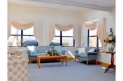 Sunlit sitting room with light blue sofas and armchairs arranged around a wooden coffee table beneath three draped windows.