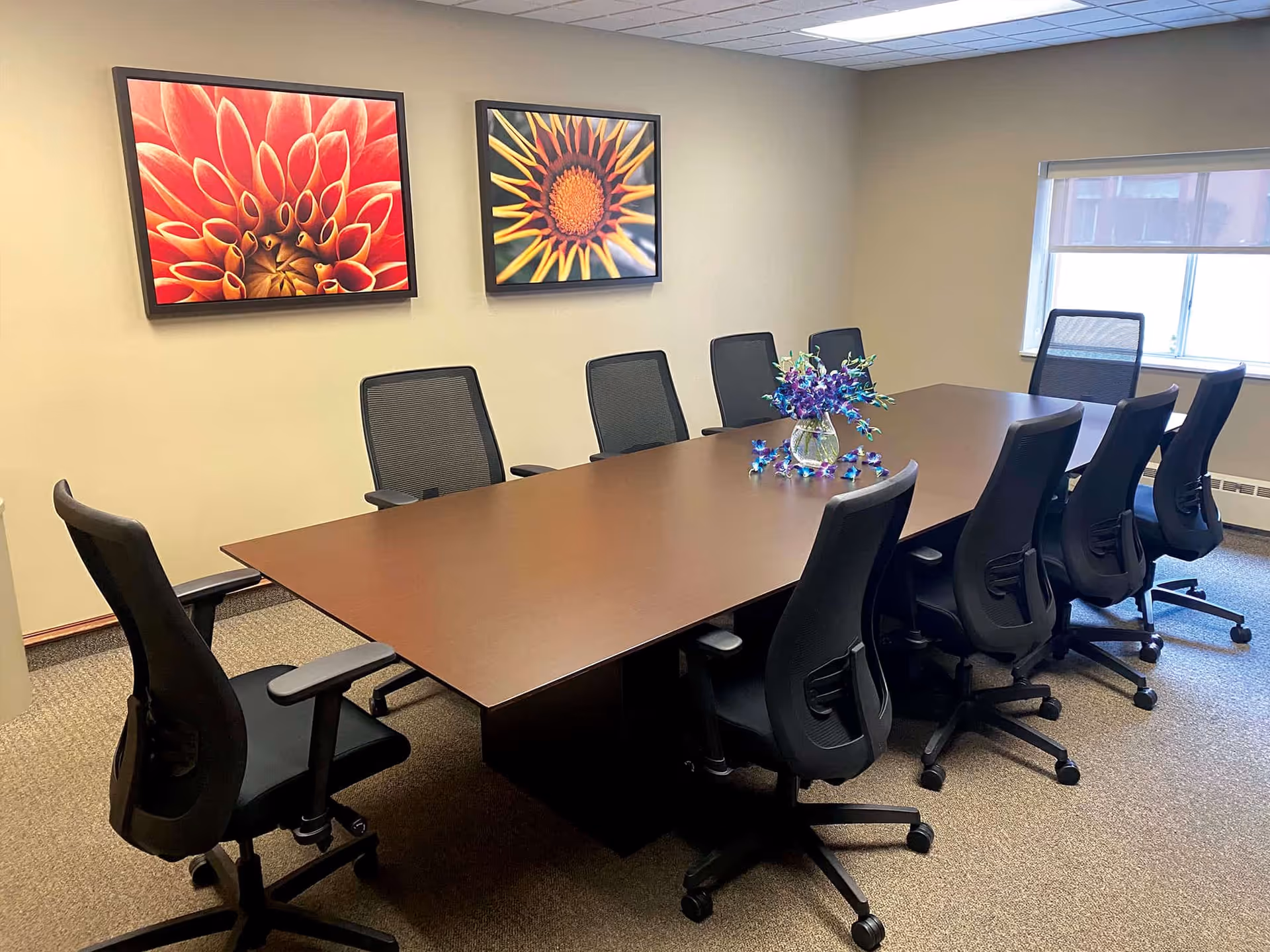 A conference-style meeting room with a long wooden table surrounded by office chairs, a floral centerpiece, and large flower artwork on the wall.