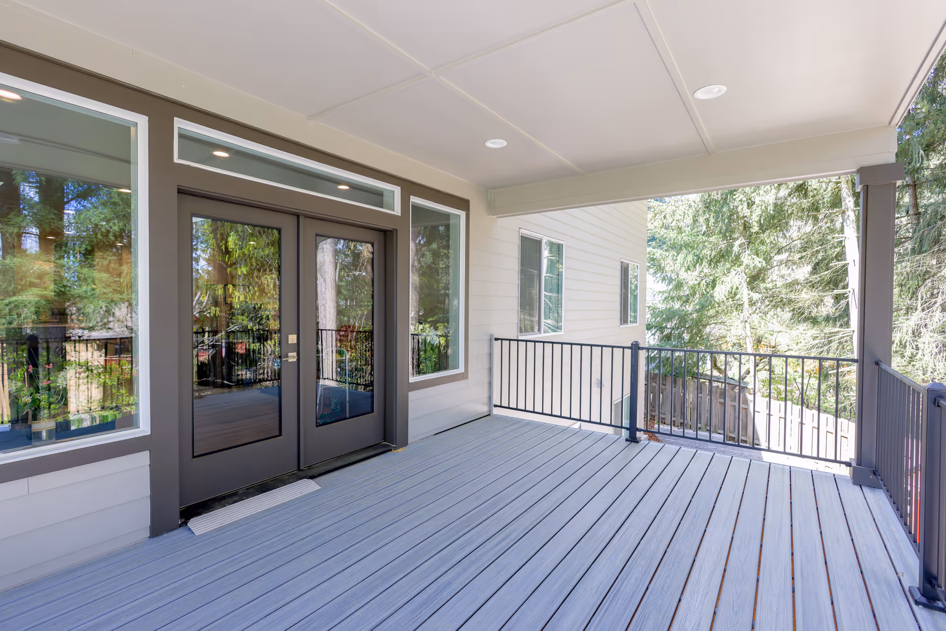 Covered outdoor deck with gray composite flooring, black metal railings, and glass double doors leading into the house overlooking trees.