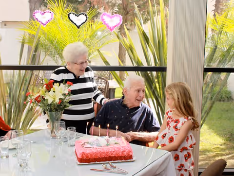 Three generations — an elderly couple and a young girl — gathered around a table with a pink birthday cake in a sunlit dining area.