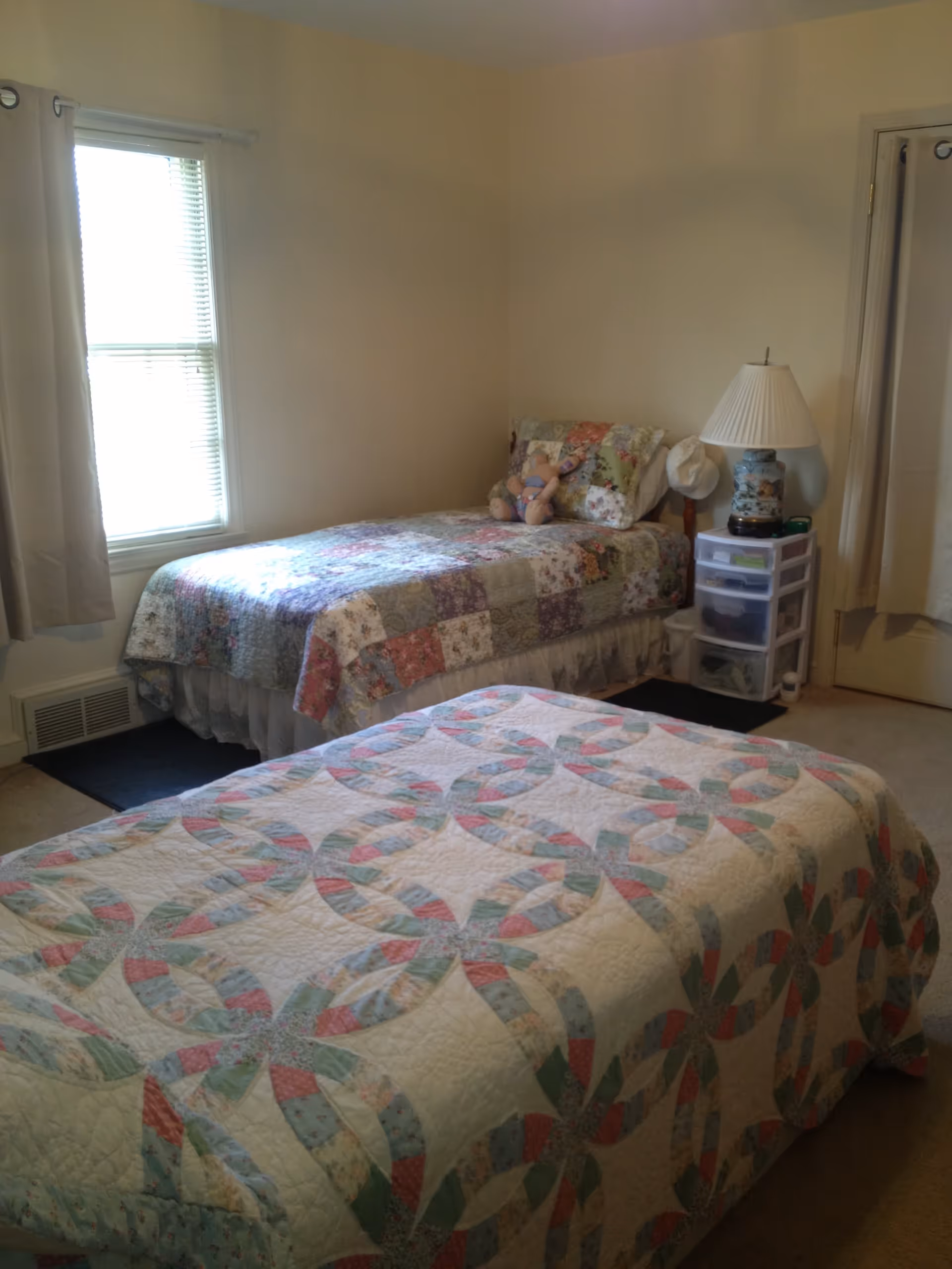 A bedroom with two twin beds covered in patchwork quilts. One bed is positioned against a wall with a window and the other bed is in the foreground. There is a small plastic drawer unit with a lamp and some items on top next to the bed against the wall. The room has beige walls and carpeted floor.
