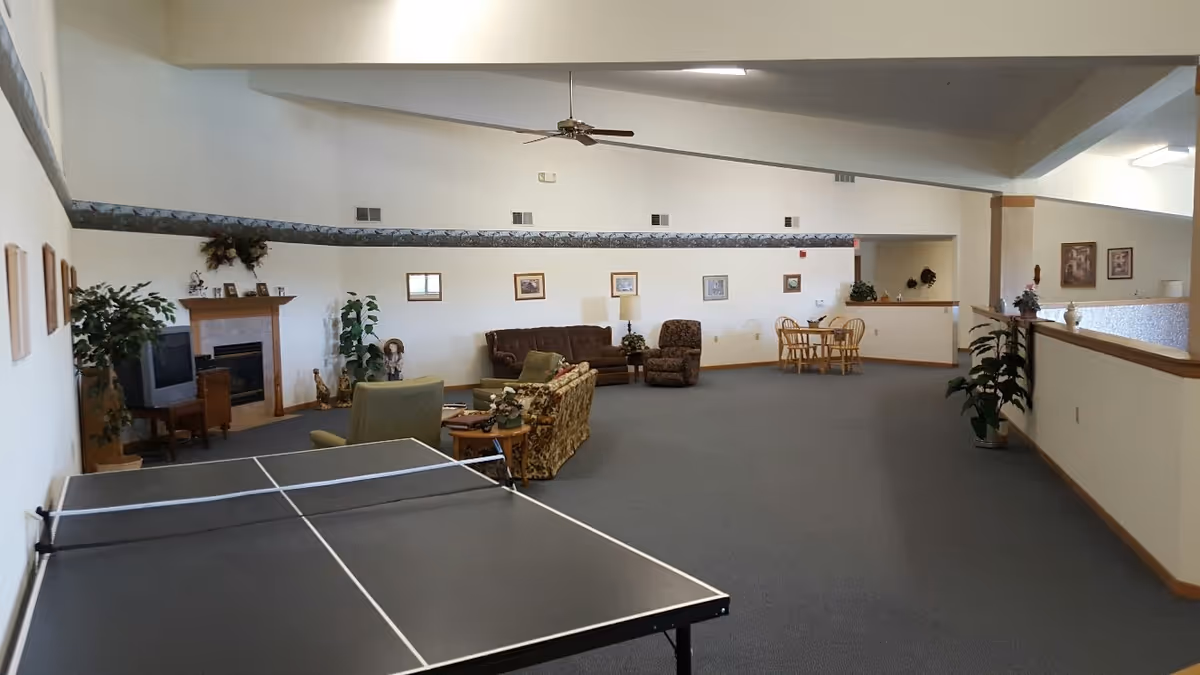 A spacious common area with a ping pong table in the foreground, several seating arrangements including sofas and armchairs, a fireplace with a TV beside it, framed pictures on the walls, and a small dining table with chairs in the background. The room has high ceilings with a ceiling fan and is decorated with plants and lamps.