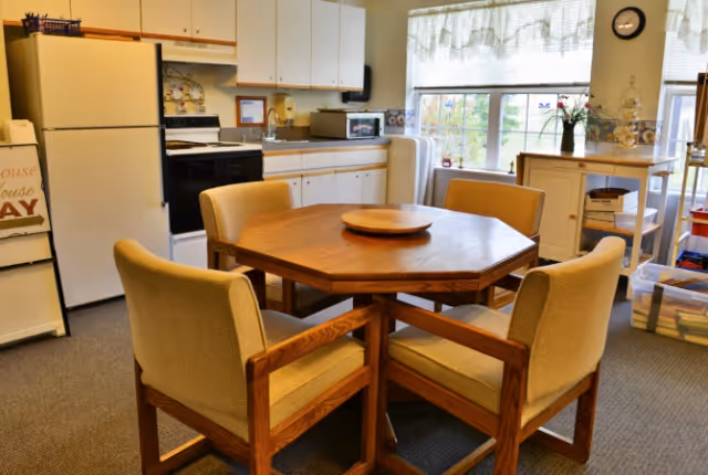 A cozy kitchen and dining area with a wooden octagonal table surrounded by four cushioned chairs. The kitchen features white cabinets, a refrigerator, a stove, a microwave, and a large window with sheer curtains letting in natural light. Various kitchen items and decorations are visible on the counters and shelves.