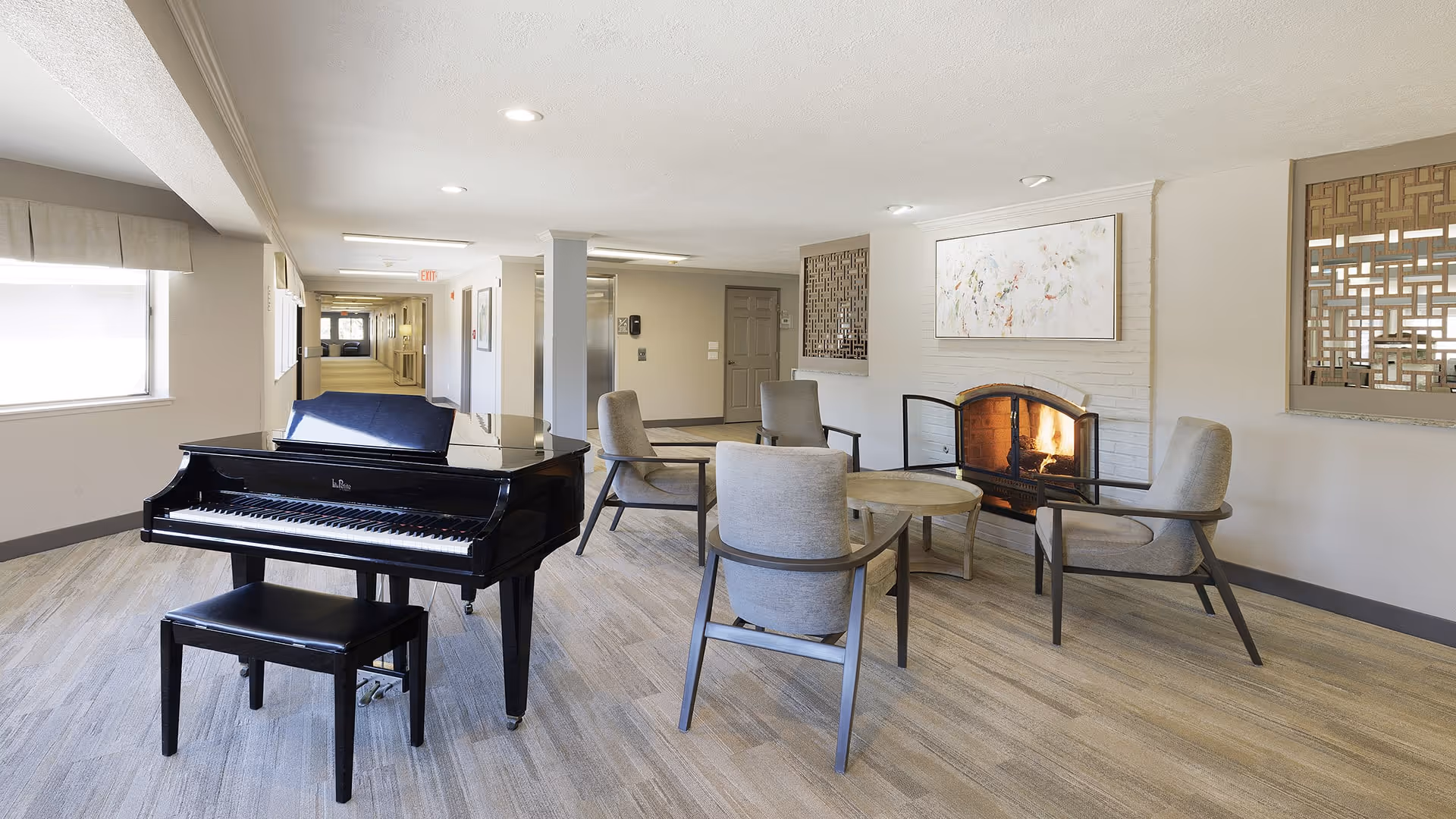 A bright common area with a black grand piano and matching bench on the left. Four gray upholstered chairs are arranged around a round wooden coffee table in front of a lit fireplace with a white brick surround. The room has light wood flooring, neutral-colored walls, and decorative wall panels. A long hallway extends in the background.