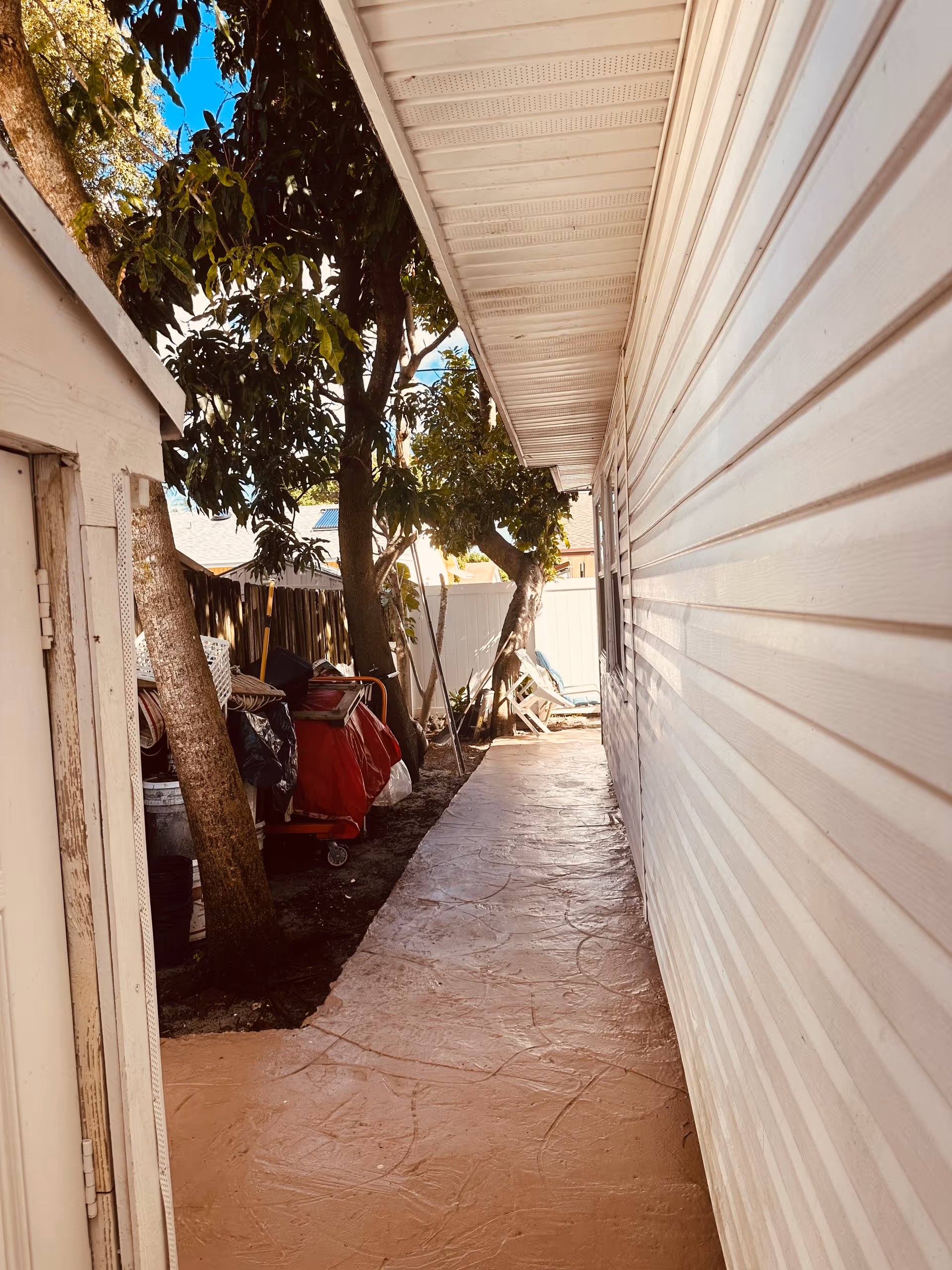 Narrow outdoor walkway beside a building with white siding, bordered by trees and a wooden fence. The walkway is paved with a textured surface and leads to a white fence at the end. Various items including a red cart and other objects are visible along the fence side.