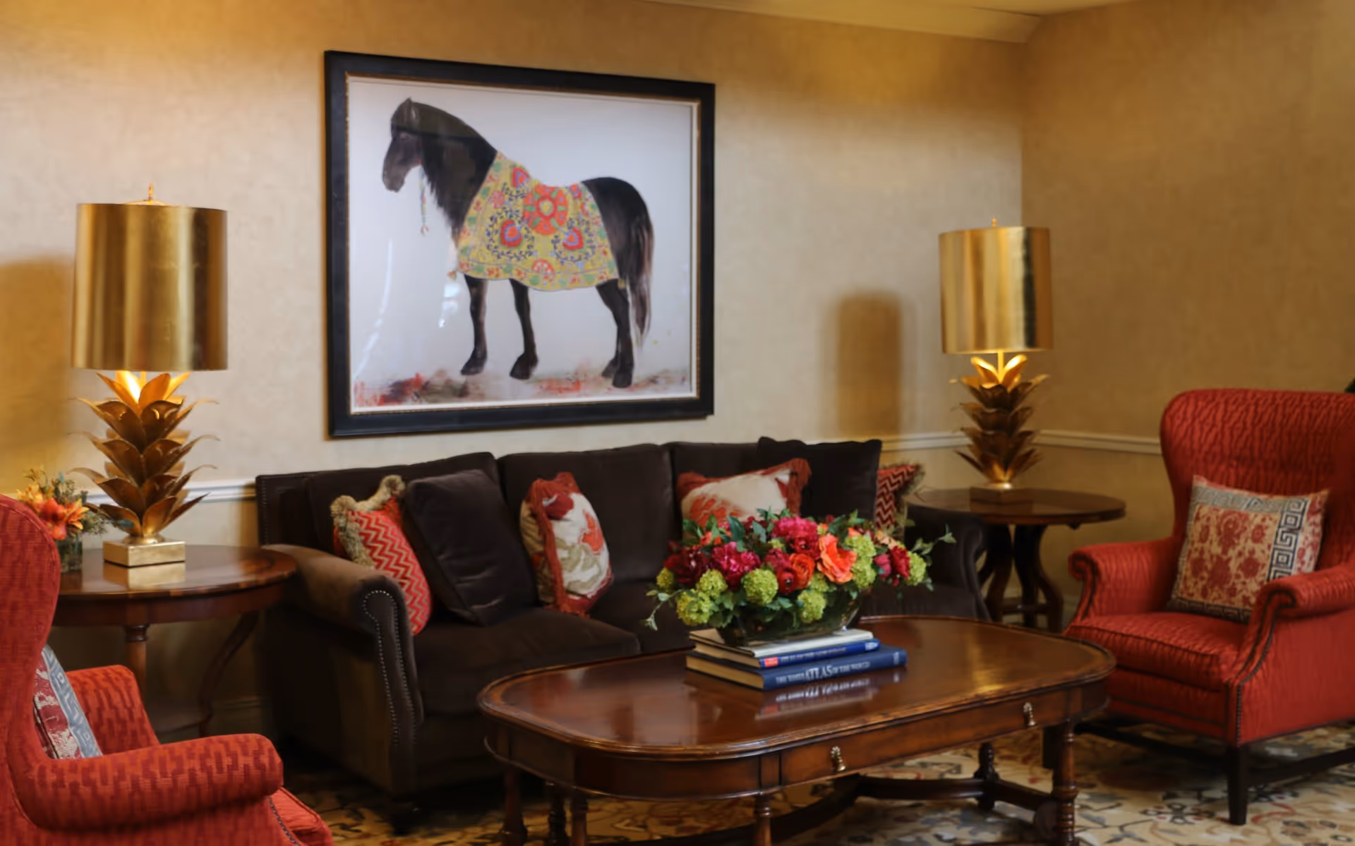 A cozy living room with a dark brown sofa adorned with multiple decorative pillows, two red armchairs with patterned cushions, a wooden coffee table with a floral arrangement and stacked books, two side tables each with a gold pineapple-shaped lamp, and a framed picture of a horse wearing a colorful blanket hanging on the beige wall.