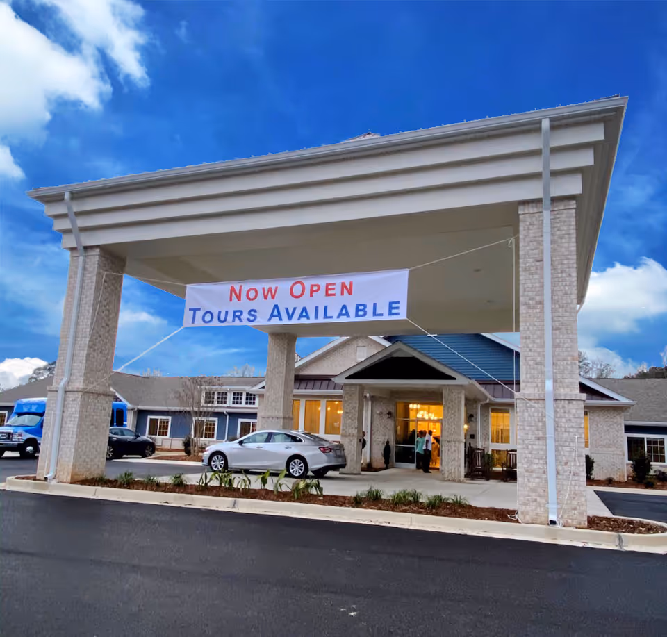Entrance of The Colonial at Old Camden facility with a large covered driveway. A banner hanging from the roof reads 'Now Open Tours Available'. Several cars are parked near the entrance, and a few people are visible near the doorway. The sky is blue with some clouds.