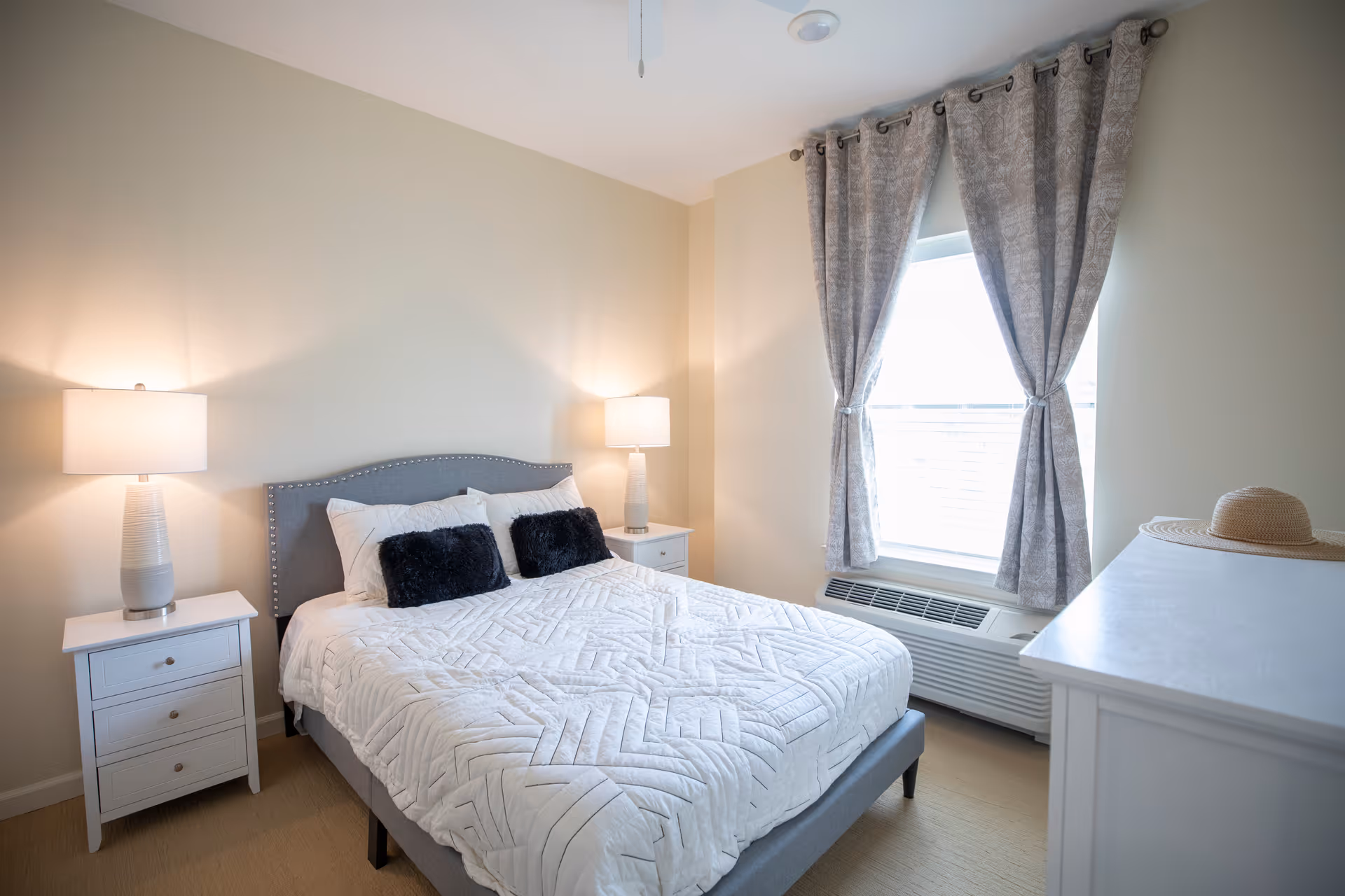 A bright bedroom with a neatly made bed featuring a white quilted bedspread and two black accent pillows. The bed has a gray upholstered headboard with nailhead trim. On either side of the bed are white nightstands with drawers, each topped with a white table lamp. A window with patterned gray curtains tied back allows natural light into the room. A white dresser with a straw hat on top is positioned near the window.