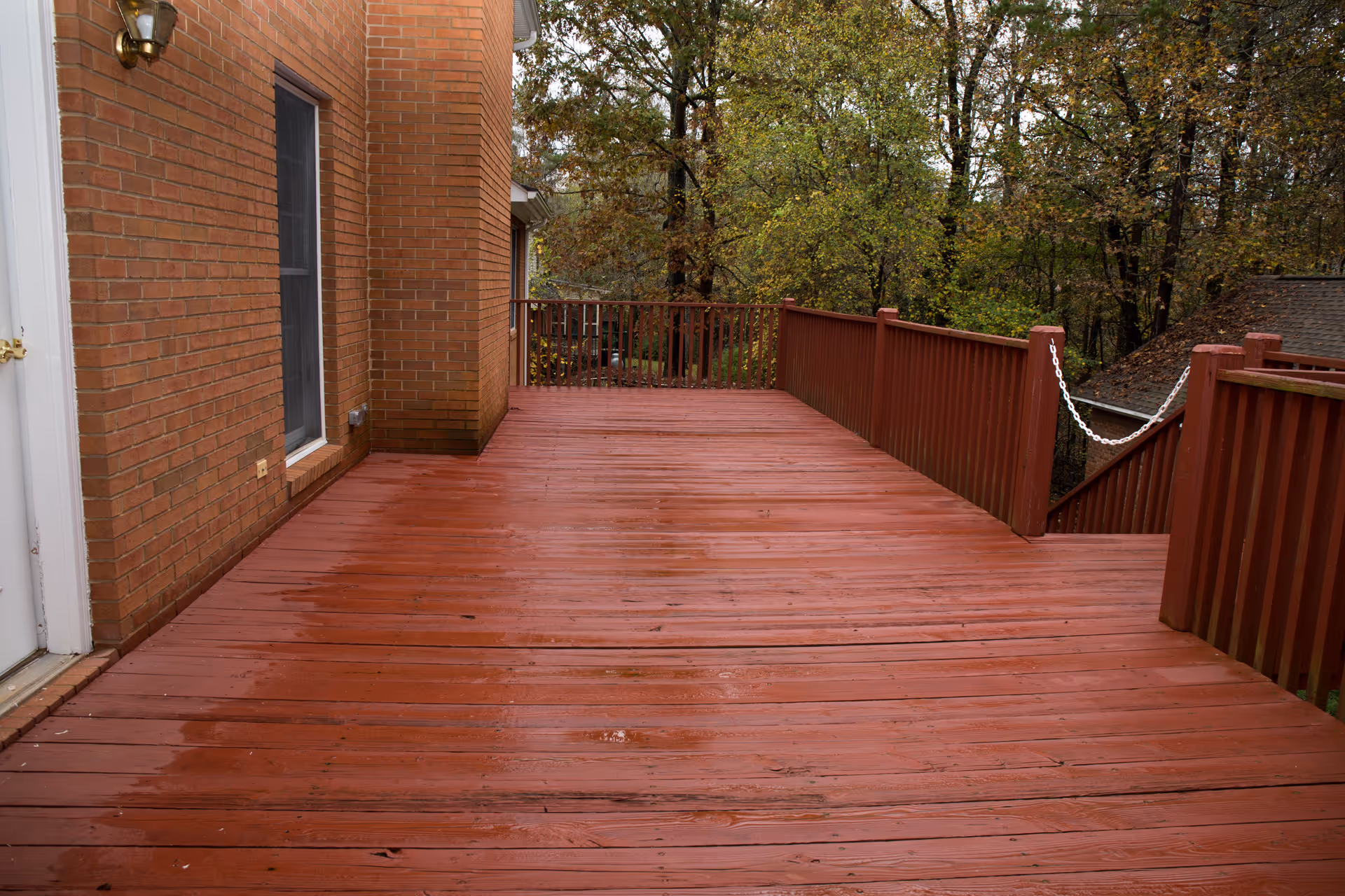 A spacious wooden deck attached to a brick building, surrounded by a red wooden railing. Trees with autumn foliage are visible in the background.