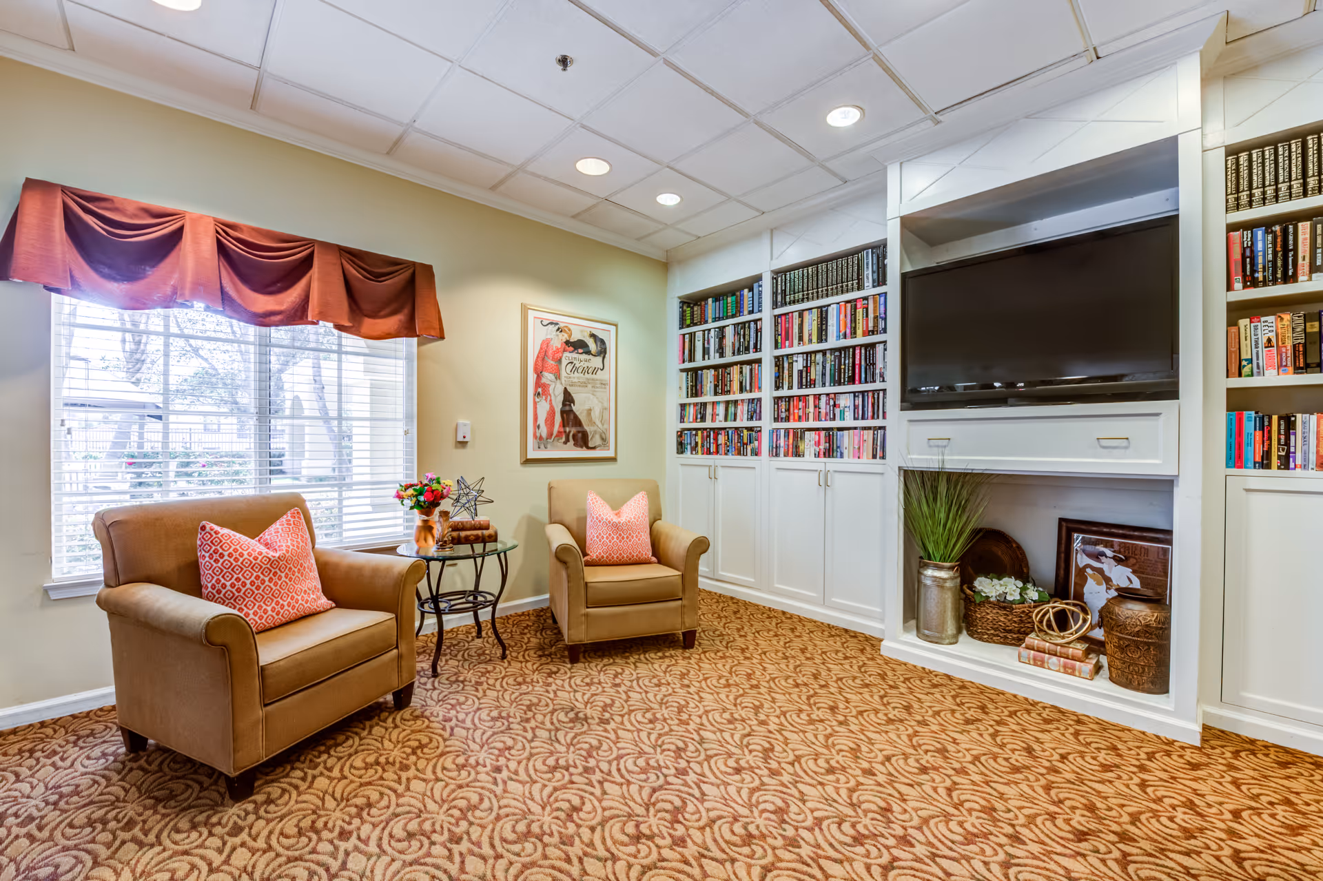 Cozy senior living common area with two brown armchairs each with a pink patterned pillow, a small round glass table with a flower arrangement, large window with red valance, built-in white bookshelves filled with books, and a flat-screen TV mounted above decorative items on the shelves.