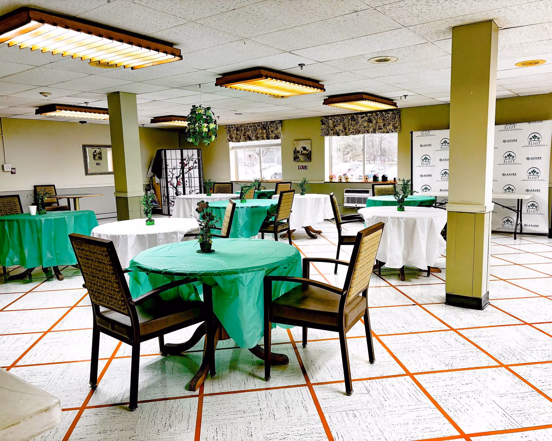 A dining area with several round tables covered with green and white tablecloths, each table having two chairs and a small plant centerpiece. The room has large windows with floral valances, fluorescent ceiling lights, and a tiled floor with a grid pattern. There is a decorative folding screen and a backdrop with the Eliot Center for Health and Rehabilitation logo in the background.