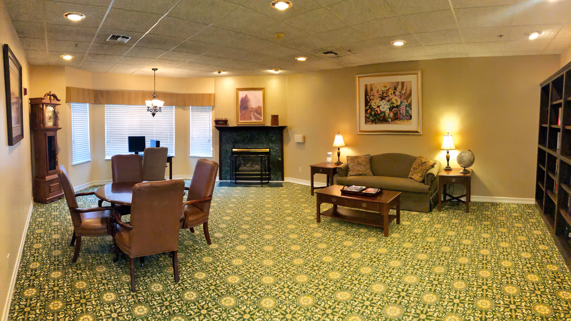 A spacious senior living lounge with sofa, chairs, fireplace, and patterned carpet.