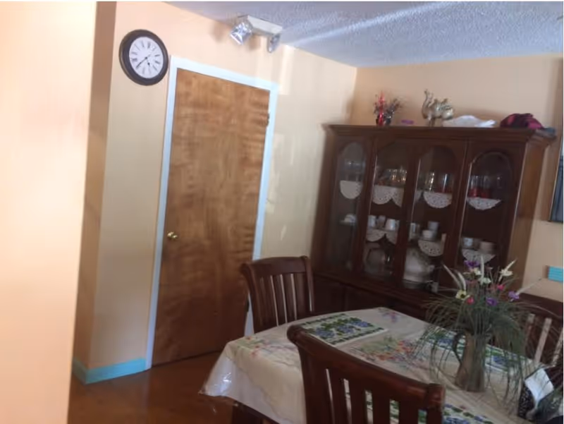 A dining room with a wooden dining table covered with a floral tablecloth and surrounded by wooden chairs. A vase with flowers is placed on the table. In the background, there is a wooden china cabinet displaying dishes and glassware. A wooden door and a wall clock are visible on the left side of the image.