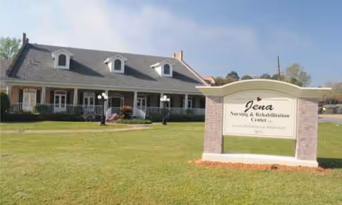 Long front view of Jena Nursing & Rehabilitation Center with a manicured lawn and sign.