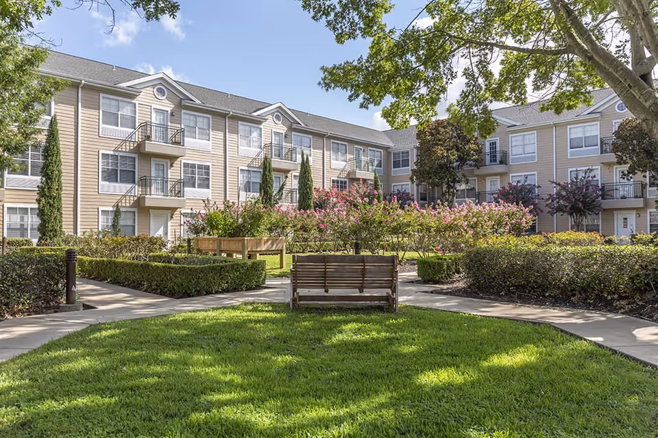 Outdoor courtyard area of a senior living facility with green grass, trimmed hedges, flowering bushes, and a wooden bench under a large tree. The background shows a three-story beige building with balconies and multiple windows under a blue sky with some clouds.