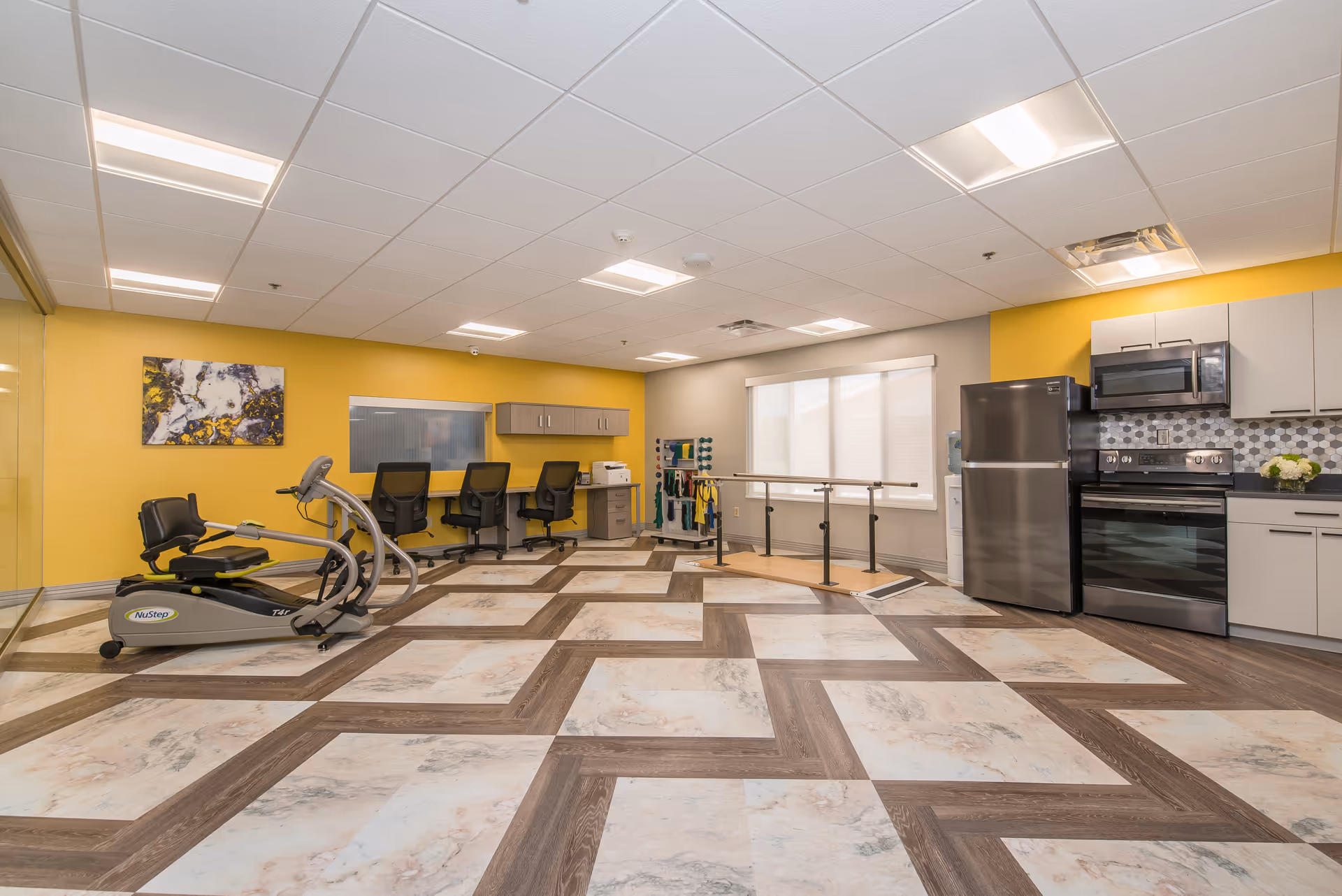 A spacious room with a patterned floor featuring a mix of wood and marble-like tiles. The room includes a NuStep exercise machine, three black office chairs at a counter with cabinets above, a set of parallel bars for physical therapy, and a modern kitchen area with a refrigerator, stove, microwave, and cabinets. The walls are painted yellow and gray, with a large window covered by blinds and a colorful abstract painting on the yellow wall.