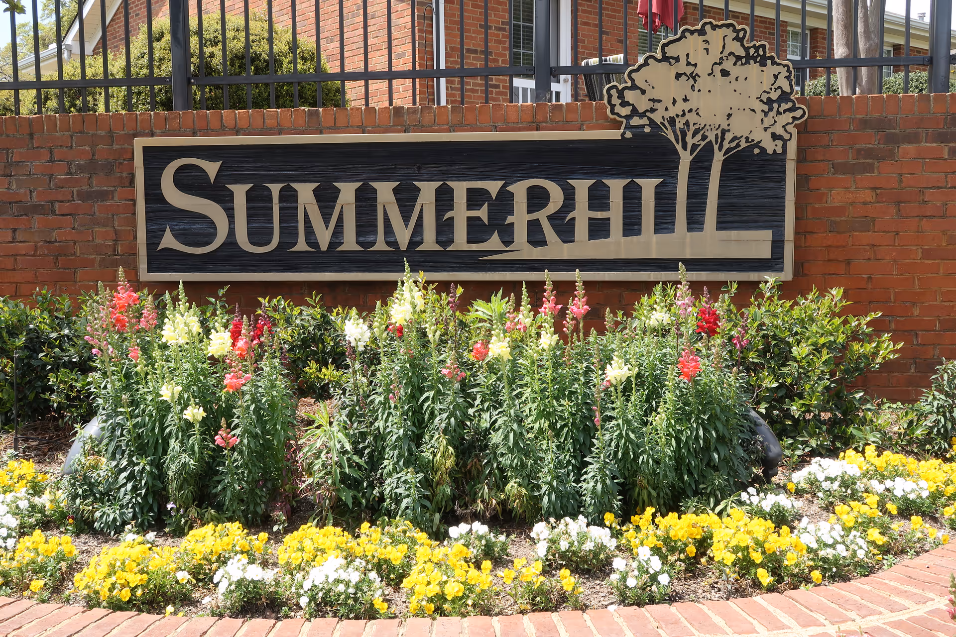 A large wooden sign reading "SUMMERHILL" mounted on a brick wall above a flowerbed with colorful flowers.