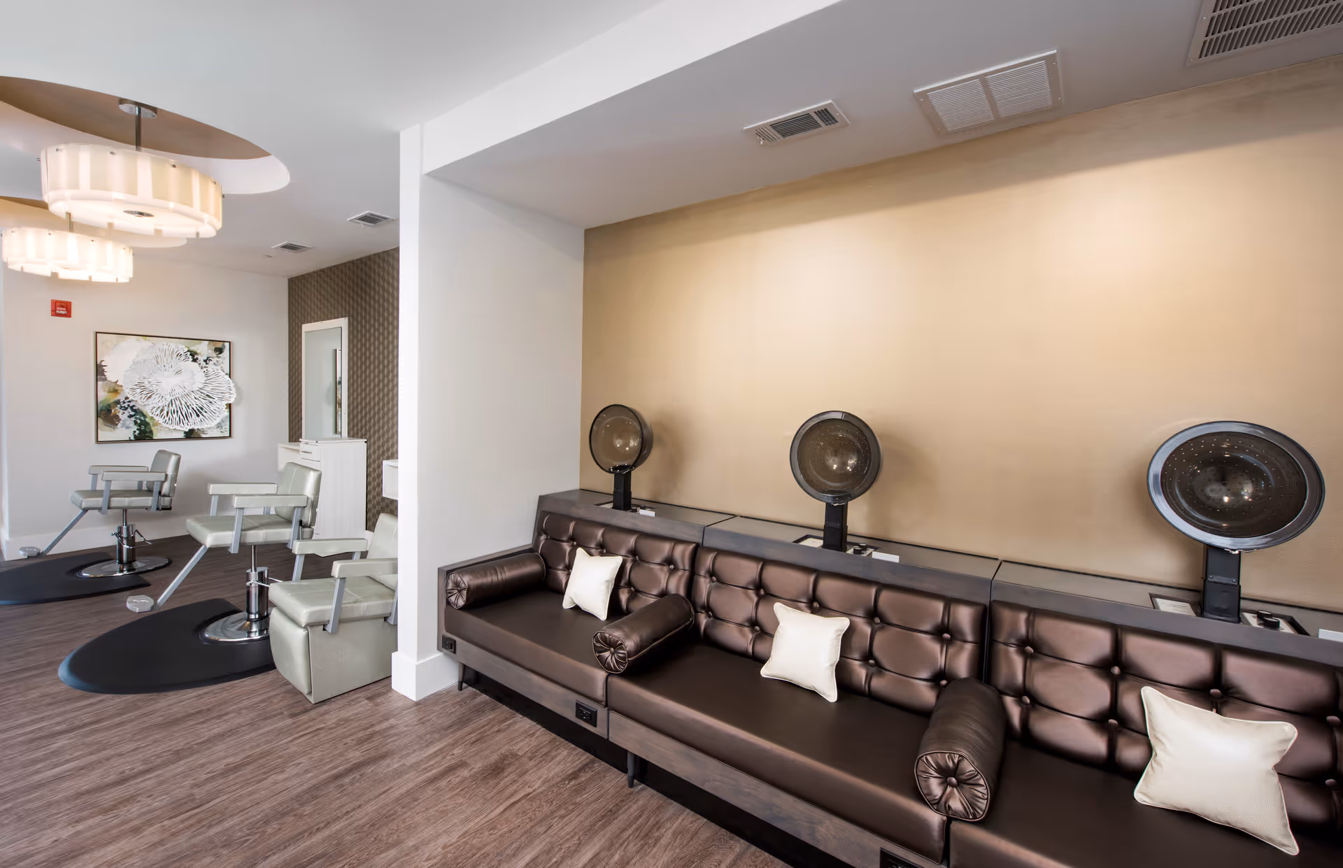 Interior view of a salon area in a senior living facility featuring three hair drying stations with brown leather cushioned seating and white pillows. To the left, there are three salon chairs with mirrors and a piece of wall art. The floor is wood, and the walls are painted in neutral tones with modern light fixtures on the ceiling.