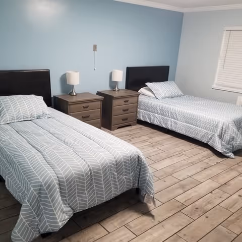 Two twin beds with matching blue patterned bedding, two nightstands with lamps, and wood-look flooring in a light blue-walled room.