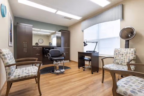 A well-lit room with wooden flooring featuring a salon chair in front of a large mirror and dark wood cabinetry. There are three patterned armchairs arranged around the room, a small desk with a lamp near a window with blinds, and a vintage hair dryer on a stand in the corner.