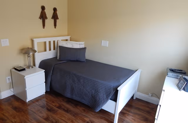 A simple bedroom with a single white bed covered with a dark gray quilt and two pillows. Next to the bed is a white nightstand with a silver lamp and a small book. The room has wooden flooring and beige walls with two decorative wooden wall hangings above the bed. On the right side, there is a white dresser with a framed photo and a telephone on top.
