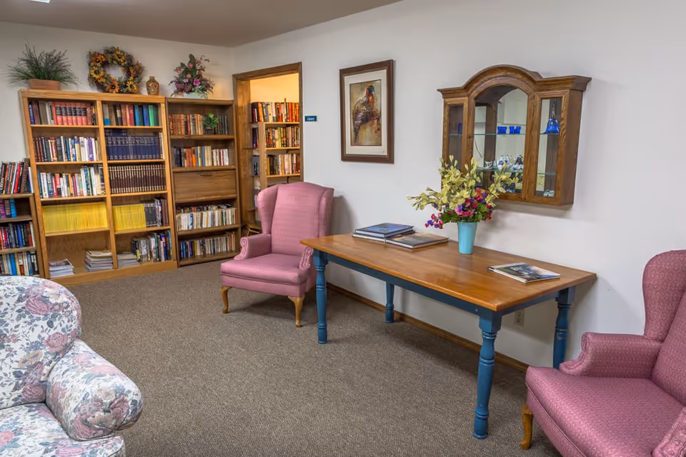 A cozy library room with wooden bookshelves filled with books, two pink upholstered armchairs, a wooden table with blue legs holding books and a vase of flowers, a floral patterned sofa partially visible, and a framed painting on the wall. A wooden cabinet with glass doors is mounted on the wall above the table.